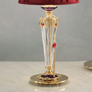 The Classic Style Swarovski® Crystal Table Lamp is an ornate piece featuring a clear, fluted column base adorned with red and purple gemstones, set on a glossy, round base. This lamp showcases an intricate design and is topped with a rich, red fabric shade. It is beautifully displayed against a soft grey wall.