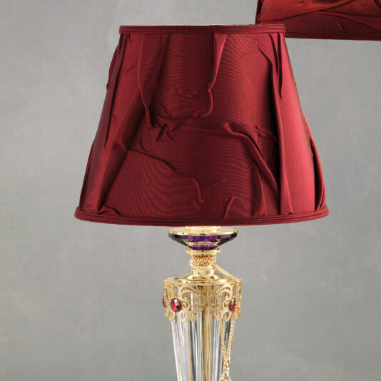 The Classic Style Swarovski® Crystal Table Lamp boasts a luxurious red, crinkled fabric lampshade and ornate gold detailing on the base, adorned with intricate designs and small Swarovski® crystal gems, adding an elegant touch. This striking piece stands against a plain, light grey wall.