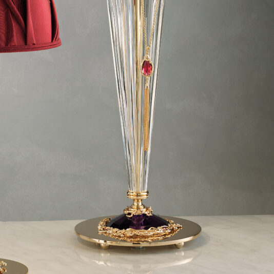 The Classic Style Swarovski® Crystal Table Lamp boasts a gold and crystal base with intricate gold embellishments and a hanging red gem. The lamp stand features vertical ridges, rests on a marble surface, and showcases the elegance of genuine Swarovski® Crystal. A portion of its red lampshade is visible on the left side of the image.