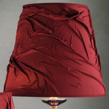 The Classic Style Swarovski® Crystal Table Lamp boasts a deep burgundy fabric lampshade with an elegant, wrinkled design. Its base is adorned with Swarovski® crystal and gold accents, adding a touch of sophistication to its overall appearance, set against a plain, neutral gray background.