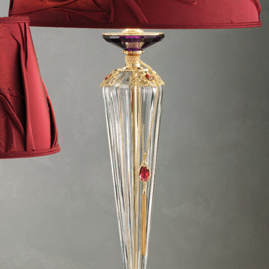 The Classic Style Swarovski® Crystal Table Lamp boasts a decorative, classic design with a clear, intricately fashioned glass base adorned with gold accents and a hanging red gemstone. Enhanced with genuine Swarovski® crystals, it features a deep red fabric lampshade with folded, ornate detailing against a muted grey background.