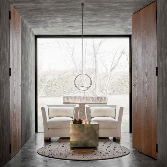 A minimalist room with concrete walls and a large window features two white armchairs facing each other. A square metallic coffee table and the Contemporary Round Pendant Light With Marble Effect are centered between the chairs. The floor has a round, decorative rug with intricate patterns.