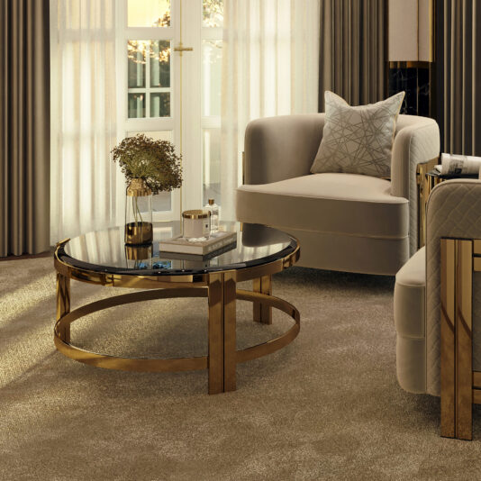 A luxurious living room features the Contemporary Round Smoked Glass Coffee Table with gold legs, accompanied by a vase of dried flowers and a few items. Two elegant upholstered armchairs rest on a soft beige carpet, while large windows allow gentle natural light to flood the space.