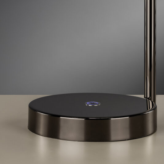 A close-up photo showcases the base of the Contemporary Round Table Lamp With Marble Effect, highlighting a circular touch sensor at its center. The sleek, modern black metal lamp rests on a smooth, beige surface and is set against a neutral gray background.