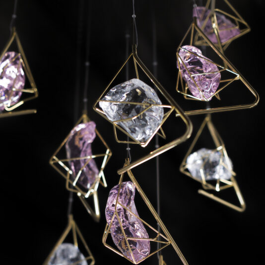 Crystal Rock Pendant Light Cluster Abstract crystal ornaments hang from thin wires against a black background. The crystals, reminiscent of those in the Crystal Rock Pendant Light Cluster, are a mix of purple and clear, held within geometric gold wire frames, creating a modern and elegant display.