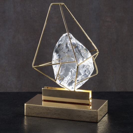 Crystal Rock Table Lamp The Crystal Rock Table Lamp is a modern sculpture that features a large, rough crystal rock encased within a geometric, gold metal frame. The crystal appears to be suspended in mid-air, complemented by the gold frame and base, all set against a matte, dark gray background.