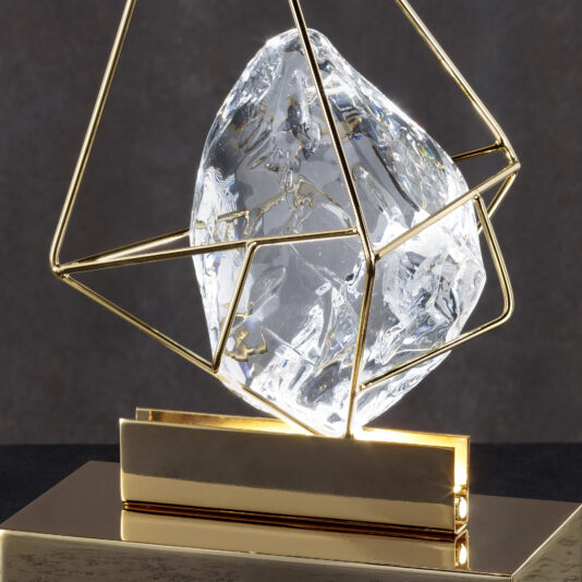 Crystal Rock Table Lamp A close-up of the Crystal Rock Table Lamp showcases its elegant, geometric design. The crystal is encased in a golden wireframe structure and stands on a glossy, gold rectangular base. The soft, dark gradient background highlights the intricate and transparent facets of the crystal.