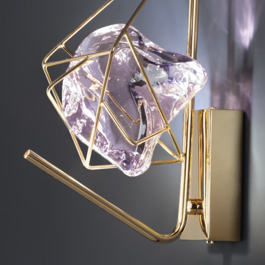 The Crystal Rock Wall Light features a modern design with a golden geometric frame encasing a sculpted, translucent purple glass piece. This fixture is mounted on a sleek, rectangular gold base against a gray wall, seamlessly blending contemporary art with functional lighting.