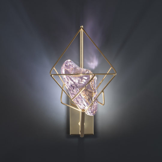 The Crystal Rock Wall Light showcases a contemporary design with a striking geometric gold frame enveloping a sizable, irregular translucent crystal. The light shines through the structure, creating complex shadows against a dark grey background.