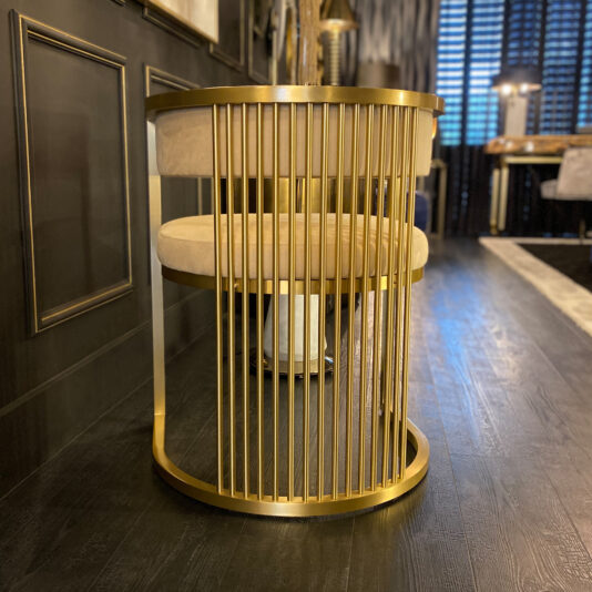 The Exclusive Contemporary High End Dining Chair boasts a chic, gold metallic frame with elegant vertical bars. This one-of-a-kind dining chair showcases a circular design and a plush, cream-colored fabric seat. It is perfectly placed in a stylish room featuring dark hardwood flooring and contemporary decor, embodying high-end aesthetics.