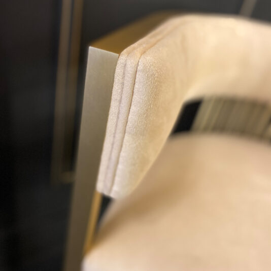 Close-up of the Exclusive Contemporary High End Dining Chair, featuring luxurious beige upholstery and a sleek metallic gold frame. The image highlights the upper corner of the chair's backrest, showcasing its soft fabric and smooth metallic finish. The blurred background accentuates the chair’s modern design.