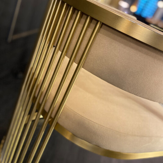 Close-up of the Exclusive Contemporary High End Dining Chair, which features a circular, cushioned seat in light beige fabric. This high-end dining chair boasts a sleek gold-colored metal frame with vertical rods for support and a shiny metallic sheen. The dark floor beneath adds striking contrast to the stool.