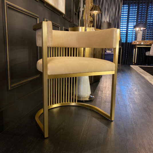 The Exclusive Contemporary High-End Dining Chair, featuring sleek beige upholstery, stylish brass accents, and an arched metal base, is positioned in an elegantly decorated room with dark wooden flooring and a dark wall panel behind it. A table and a window with closed blinds are visible in the background, enhancing the sophisticated modern ambiance.