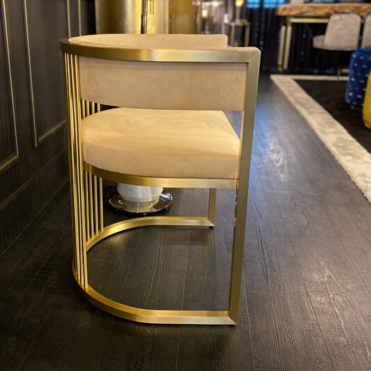The Exclusive Contemporary High End Dining Chair, adorned with a plush beige seat and a stylish gold frame that includes a semi-circular backrest and vertical metal slats, is positioned elegantly on a sleek dark wooden floor. This luxurious dining chair complements the modern furniture and decorative elements in the backdrop.