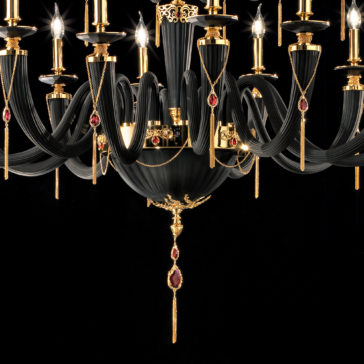Large Classic Black Chandelier With Swarovski® Crystals The Large Classic Black Chandelier With Swarovski® Crystals is an elegant piece featuring gold accents and red Swarovski® crystal details. This chandelier showcases multiple candle-like lights arranged in a circular pattern, with gold chains and ornate designs enhancing its luxurious appearance against a dark background.