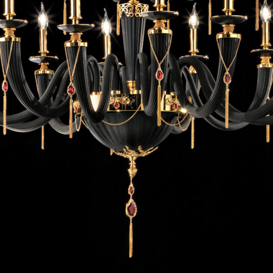 The Large Classic Black Chandelier With Swarovski® Crystals is an elegant piece featuring gold accents and red Swarovski® crystal details. This chandelier showcases multiple candle-like lights arranged in a circular pattern, with gold chains and ornate designs enhancing its luxurious appearance against a dark background.