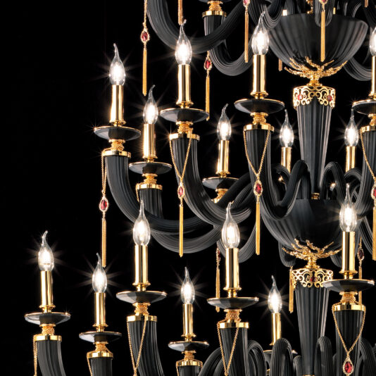 A large classic black chandelier with intricate black and gold designs, adorned with candle-shaped lights. This ornate multi-tiered piece features small red accents and sparkling Swarovski® crystals, standing out prominently against a black background.