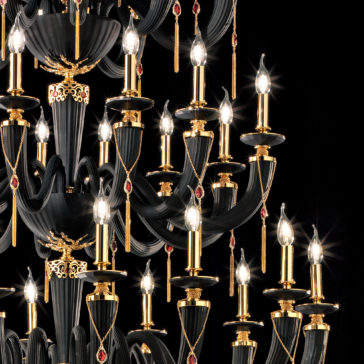 Large Classic Black Chandelier With Swarovski® Crystals A close-up of the Large Classic Black Chandelier With Swarovski® Crystals showcases numerous candle-like lights. The chandelier is adorned with gold details and sparkling Swarovski® crystals, with pendant red jewels hanging from various points. The dark background enhances the brilliance of the illuminated lights.