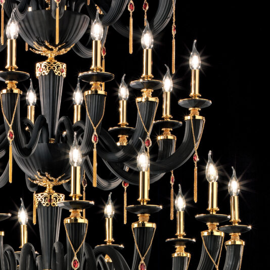 A close-up of the Large Classic Black Chandelier With Swarovski® Crystals showcases numerous candle-like lights. The chandelier is adorned with gold details and sparkling Swarovski® crystals, with pendant red jewels hanging from various points. The dark background enhances the brilliance of the illuminated lights.