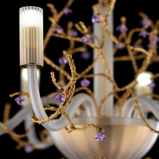 A close-up photo of the Large Modern Chandelier With Swarovski Crystal Flowers reveals its detailed floral design. The chandelier boasts gold branches adorned with small purple flowers and translucent white lamp shades, emitting a soft glow. The black background highlights the intricate details of this modern centerpiece.
