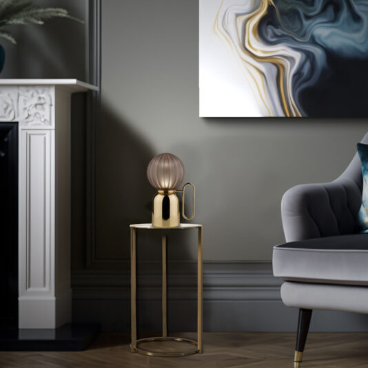 A modern living room featuring a gray armchair with wooden legs, a gold side table topped with the Small Satin Taupe Glass Cordless Battery Table Lamp, and abstract wall art with swirling black, white, and gold patterns. A fireplace with intricate detailing is partially visible on the left.