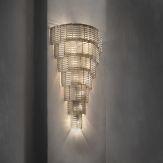 Oversized Tiered Murano Glass Wall Light The Oversized Tiered Murano Glass Wall Light features a modern chandelier design with tiered glass pieces arranged in a conical shape and mounted on a wall with a soft grey finish. It emits a warm glow, creating an elegant and decorative effect.