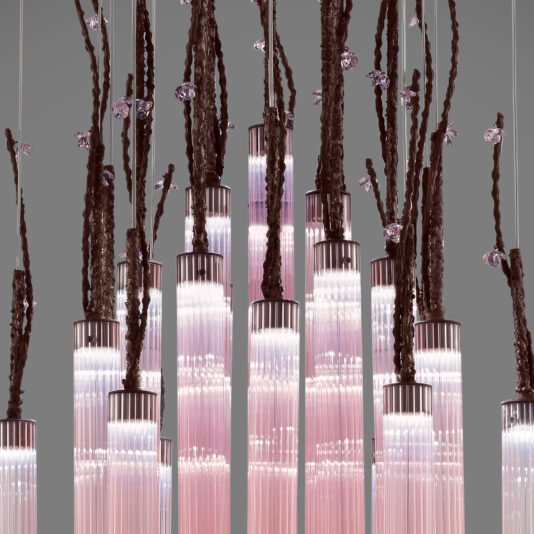 The Pink Fluted Glass Chandelier With Swarovski Crystal Flowers showcases long, cylindrical light fixtures with a gradient from pale pink to clear at the bottom. Organic, branch-like structures with a dark brown texture extend upwards, some adorned with sparkling Swarovski crystal flowers against a gray background.