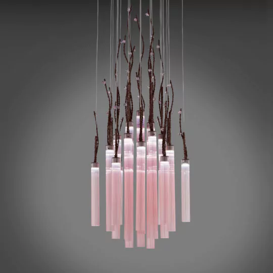 The Pink Fluted Glass Chandelier With Swarovski Crystal Flowers features wire suspensions that support its design. Sculpted branches are interwoven among the elongated, pink-toned glass tubes, adding a naturalistic and artistic touch to the chandelier. It is set against a neutral grey background.