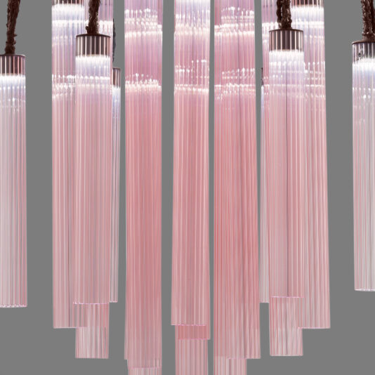 A series of vertical, cylindrical pendant lights hang from the ceiling. Each light, reminiscent of the Pink Fluted Glass Chandelier With Swarovski Crystal Flowers, features numerous translucent pink rods arranged in a circular pattern, emitting a soft, ambient glow against a neutral gray background.