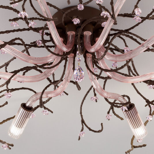 A detailed look at the Pink Glass Branch Ceiling Light With Swarovski Crystal Flowers reveals its ornate, curved glass arms designed to resemble branches, adorned with Swarovski crystal flowers and faceted hanging drops. The chandelier features lights encased in ribbed glass shades that emit a soft glow.