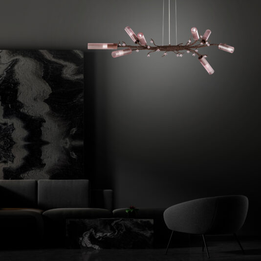 In a modern, dimly-lit living room, the Pink Glass Branch Chandelier With Swarovski Crystal Flowers hangs gracefully from the ceiling, casting a warm glow. The room features a dark grey sofa, a round grey chair, and a coffee table, all complemented by abstract, dark marbled walls.