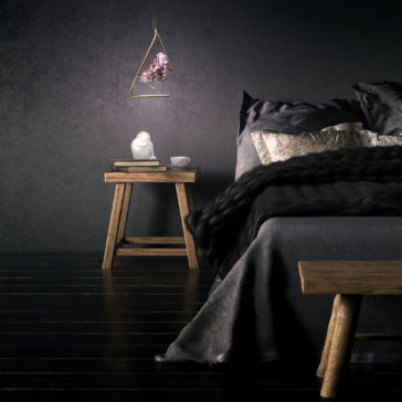 A minimalist bedroom showcases a wooden nightstand adorned with a small statue, books, and a white cup. The Rose Crystal Rock Pendant Light softly illuminates the space from above. Dark linens cover the bed, which is complemented by metallic accent pillows, all set against black walls and floors.