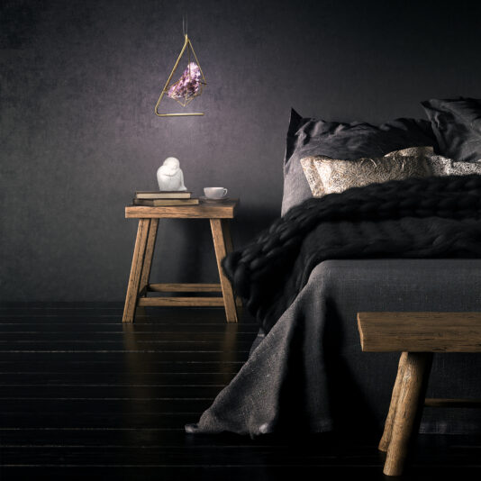 Rose Crystal Rock Pendant Light A minimalist bedroom showcases a wooden nightstand adorned with a small statue, books, and a white cup. The Rose Crystal Rock Pendant Light softly illuminates the space from above. Dark linens cover the bed, which is complemented by metallic accent pillows, all set against black walls and floors.