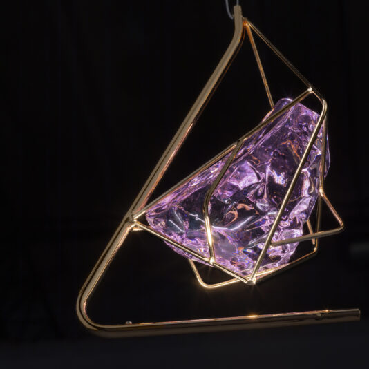 Rose Crystal Rock Pendant Light A contemporary hanging light fixture with a geometric, gold metal frame encases the stunning Rose Crystal Rock Pendant Light. When illuminated, the vibrant purple crystal casts a soft glow against the dark background, accentuating the striking design of the fixture.