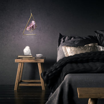 A modern bedroom with a dark aesthetic features a wooden side table next to the bed, holding a white statue, a cup, and books. Above the table hangs the Rose Crystal Rock Pendant Light with its geometric design and central rose crystal rock pendant. The bed is adorned with dark linens and decorative pillows.