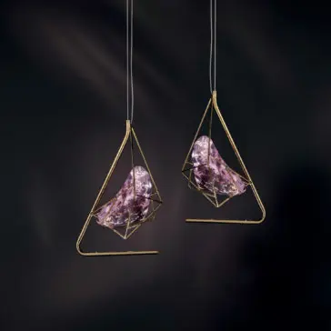 Two Rose Crystal Rock Pendant Lights, featuring gold geometric frames that encase abstract purple glass forms, are suspended against a dark backdrop, creating a modern and artistic ambiance.