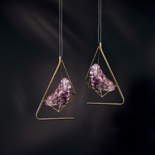 Two Rose Crystal Rock Pendant Lights, featuring gold geometric frames that encase abstract purple glass forms, are suspended against a dark backdrop, creating a modern and artistic ambiance.