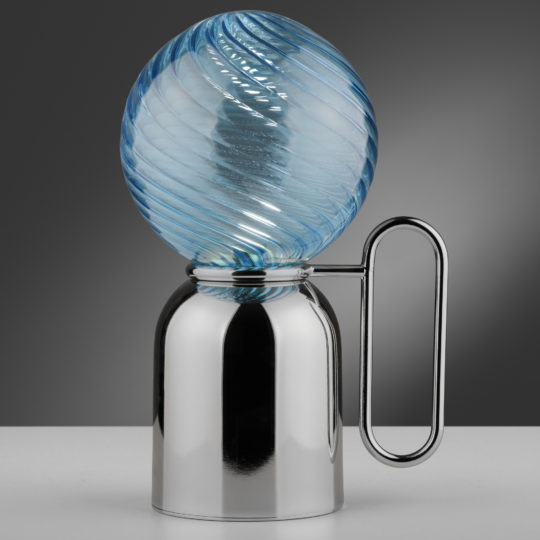 The Modern Blue Glass Cordless Battery Table Lamp is a contemporary decorative piece with a sleek metallic base, complete with a loop handle and topped by a spiraled blue glass sphere. Set against a smooth gradient from dark to light gray, it captures attention and stands out beautifully.