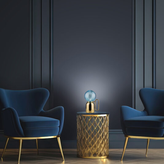 A sophisticated interior setup features two navy blue armchairs with golden legs facing each other, separated by a round gold side table. On the table is a Modern Blue Glass Cordless Battery Table Lamp with a spherical blue glass shade. The background is a dark-paneled wall.
