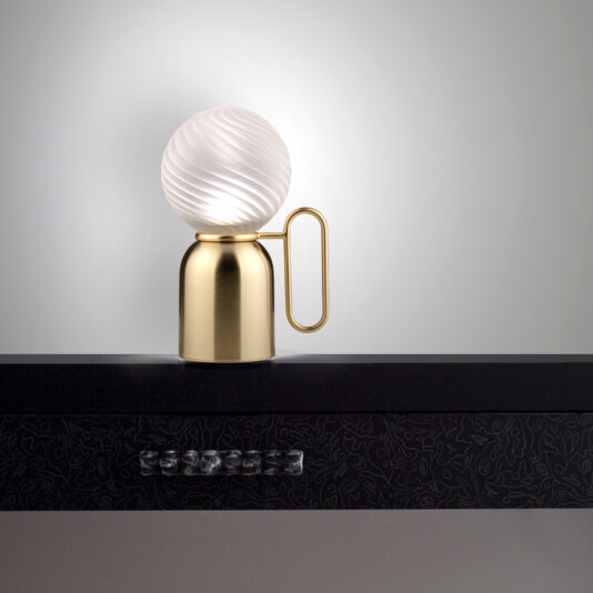 A Small Modern Glass Cordless Battery Table Lamp with a frosted, swirled glass globe and a metallic gold base sits on a dark, patterned shelf. This stylish lamp features a unique design, including a loop handle attached to the base. The background is a plain, light gray wall.