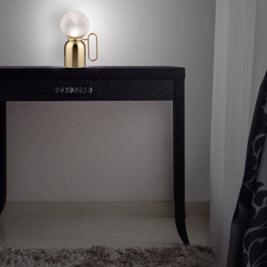 A Small Modern Glass Cordless Battery Table Lamp with a spherical, frosted glass shade and a brass base is placed on a sleek black table. The table stands against a light gray wall with a sheer curtain partially visible. A fluffy gray rug covers part of the tile floor underneath.