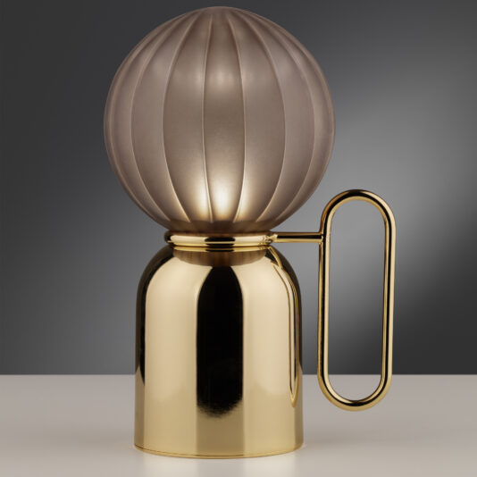 The Small Satin Taupe Glass Cordless Battery Table Lamp showcases a contemporary design with a round, frosted shade sitting on a sleek, gold-colored metallic base. The base is adorned with a unique gold handle on its side, set against a gradient background that transitions from dark to light gray.
