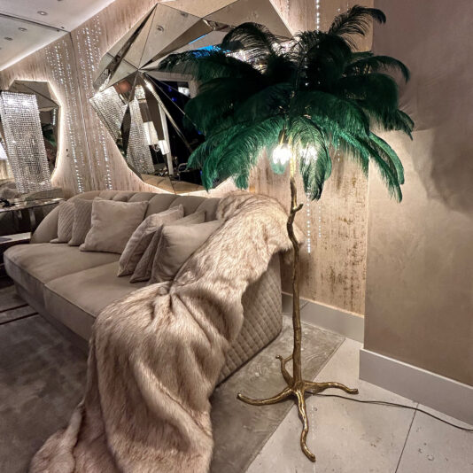 A luxurious living room features a beige sofa with plush cushions and a faux fur throw. The "Striking Palm Tree Floor Lamp With Green Feathers" provides lighting, its ornate gold design adding flair. Modern mirrored decorations adorn the walls, enhancing the space's opulent and elegant ambiance.