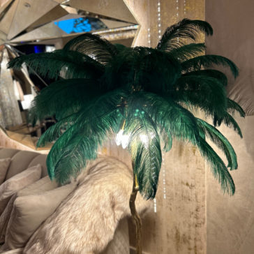 Striking Palm Tree Floor Lamp With Green Feathers A Striking Palm Tree Floor Lamp With Green Feathers is placed beside a cream-colored couch with a fur throw. The lamp is illuminated, casting a soft light in the room with mirror and metallic accents in the background.