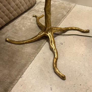 Striking Palm Tree Floor Lamp With Green Feathers A close-up of a golden three-legged table base featuring a design that mimics twisted roots or branches, set on a beige rug and marble floor. In the background, you can see part of the Striking Palm Tree Floor Lamp With Green Feathers next to a quilted fabric item.