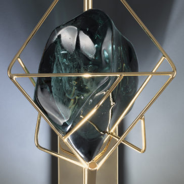 Viridian Green Crystal Rock Wall Light The Viridian Green Crystal Rock Wall Light features a sleek black stone with swirling patterns encased in a modern, geometric gold metal frame. Complemented by subtle accents of viridian green crystal, the dark glossy textures and lustrous metal create a striking contrast against a light gray background.