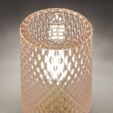 A close-up of a decorative, cylindrical, textured glass container with a honeycomb pattern. The glass is translucent, and a soft light illuminates the interior, highlighting its intricate design. Reminiscent of the Wall Light With Balloton Effect Glass Shades, the background is a neutral gray.