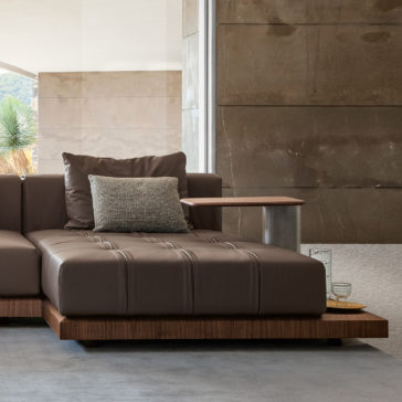 Contemporary High End Modular Corner Sofa A modern living room showcases a sleek, contemporary High-End Modular Corner Sofa with an attached wooden side table. The wall behind is clad in textured stone, with a large window allowing natural light to fill the room. A green plant is visible through the window, and there are glass items on the side table.