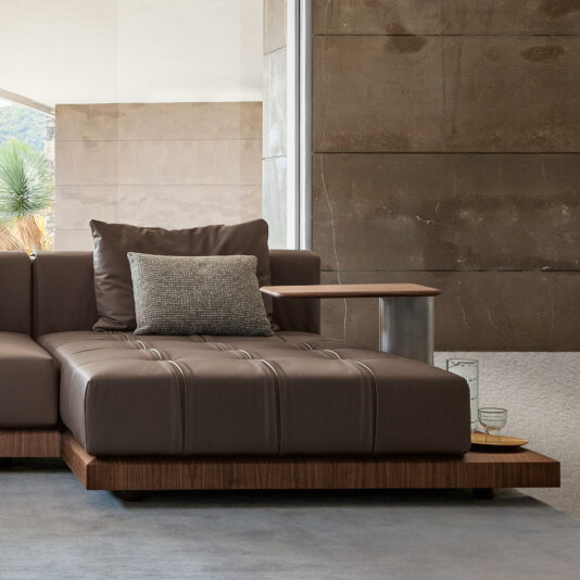 A modern living room showcases a sleek, contemporary High-End Modular Corner Sofa with an attached wooden side table. The wall behind is clad in textured stone, with a large window allowing natural light to fill the room. A green plant is visible through the window, and there are glass items on the side table.
