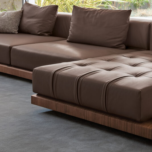 A modern living room with a Contemporary High End Modular Corner Sofa and a matching tufted ottoman on a dark gray carpet. Sunlight filters in through a window, casting a soft glow on the minimalist, stylish furniture.
